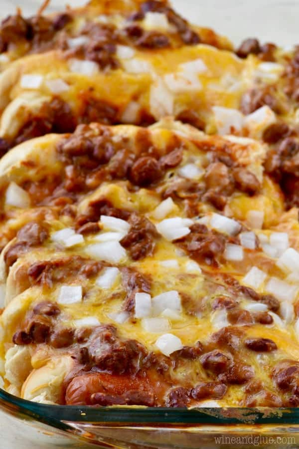 In a clear casserole dish, the Oven Baked Chili Cheese Dog is packed with cheese, chili, and diced white onions are on top. 