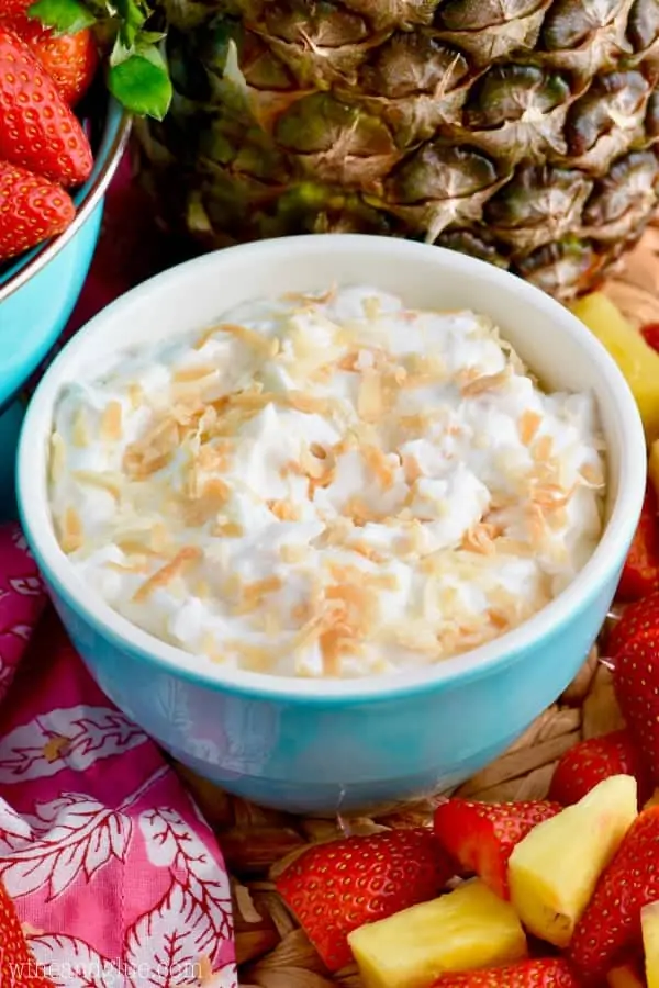 In a blue bowl, the Fruit Dip has toasted coconut flakes on top and surrounded by strawberries and pineapples. 