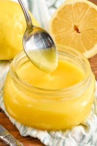 Overhead photo of a spoon dipping into jar of Lemon Curd Recipe. Lemons in the background.