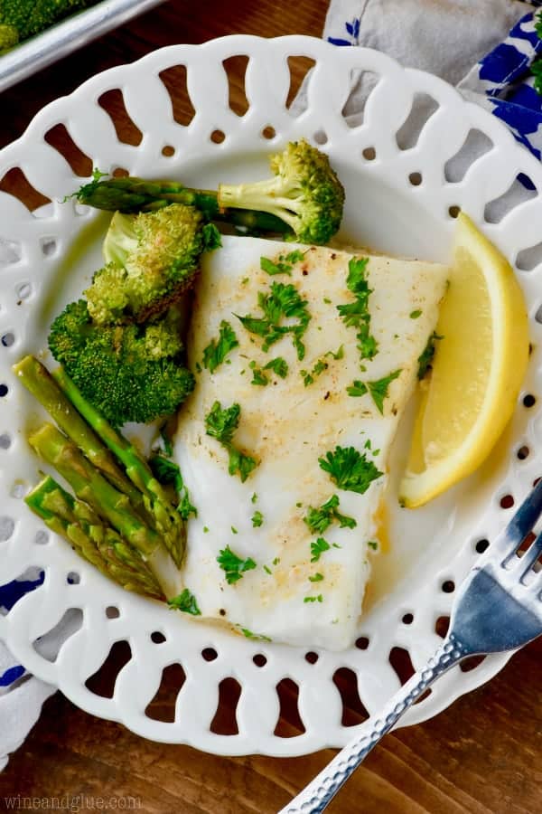 On a white plate, the Baked Cod has a golden brown sear,  topped with parsley, and surrounded with baked asparagus and broccoli. 