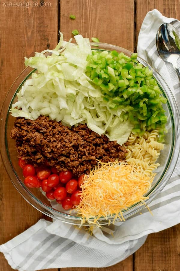 The ingredients for the Taco Pasta Salad in a clear white bowl: shredded cheese, cherry tomatoes, ground beef, lettuce, onions, pasta, and green peppers. 