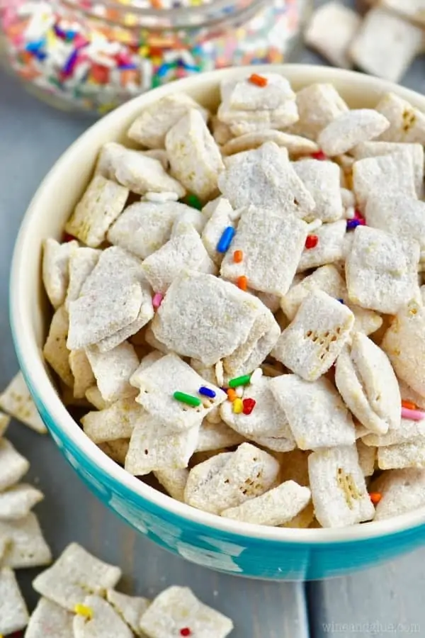 In a blue bowl, the Funfetti Cake Batter Muddy Buddies is powdered with some dry white cake batter and sprinkled with rainbow sprinkles. 