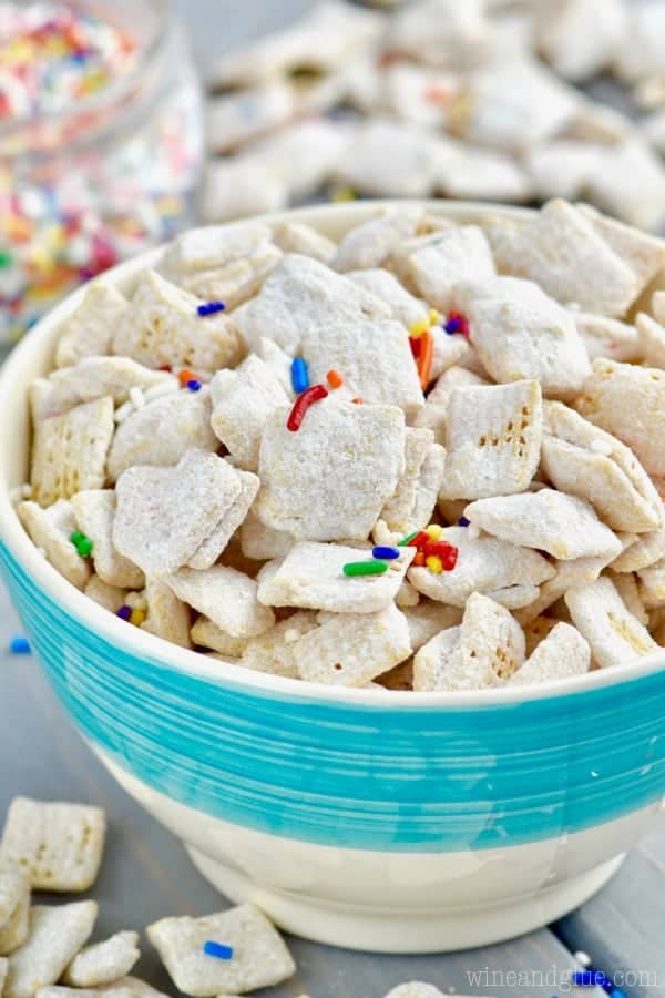 In a blue bowl, the Funfetti Cake Batter Muddy Buddies is powdered with some dry white cake batter and sprinkled with rainbow sprinkles. 