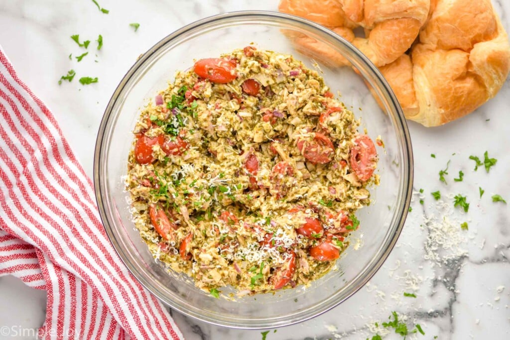 Overhead photo of a mixing bowl of ingredients combined for Pesto Chicken Salad recipe. Croissants on counter beside mixing bowl.