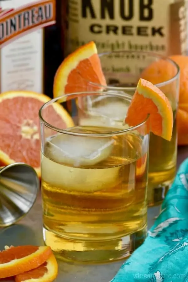 In a glass, the Desert Wasteland Whiskey Cocktail has a large cube of ice and a slice of orange on the brim of the glass. 