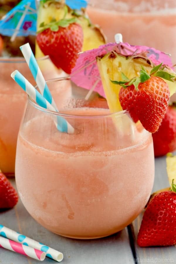 A glass of the Frozen Sangria Slush with paper umbrellas, a slice of a strawberry, and a slice of pineapple on the rim of the glass.