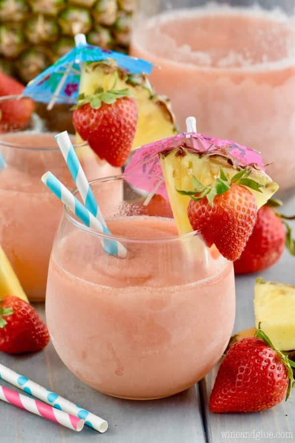 A glass of the Frozen Sangria Slush with paper umbrellas, a slice of a strawberry, and a slice of pineapple on the rim of the glass.