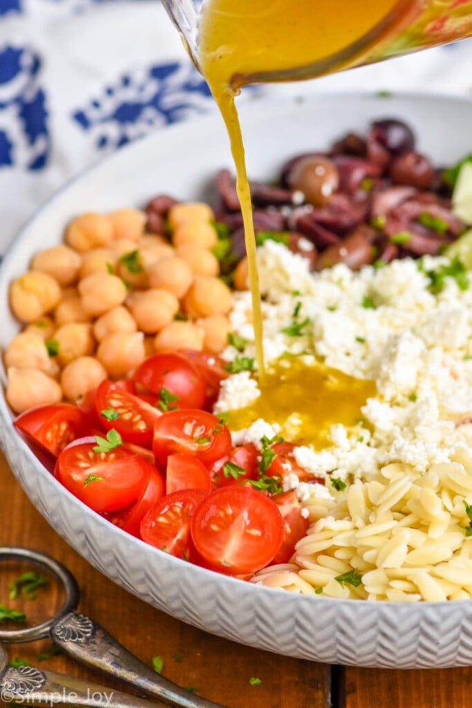 Overhead photo of a bowl of ingredients for Easy Orzo Salad recipe with dressing being poured onto ingredients.