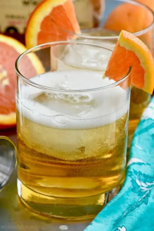 In a glass, the Desert Wasteland Whiskey Cocktail has a large cube of ice and a slice of orange on the brim of the glass. 