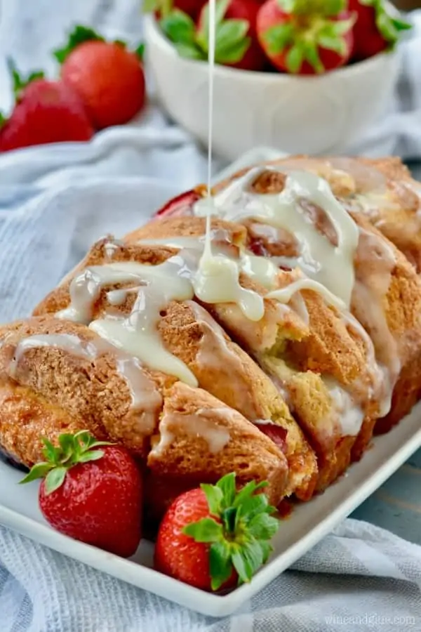 On a white plate, the Strawberries and Cream Pull Apart Bread has a beautiful golden brown crust, and some white cream is being drizzled on top of it. 