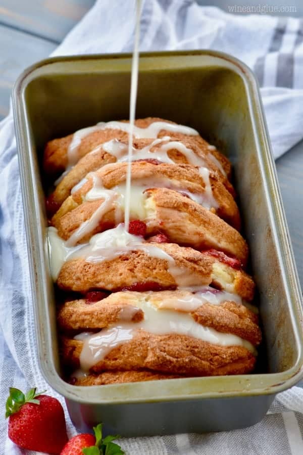 In a bread pan, the Strawberry and Cream Pull Apart Bread is fully baked with a a golden brown crust, and some white icing is being drizzled on top. 
