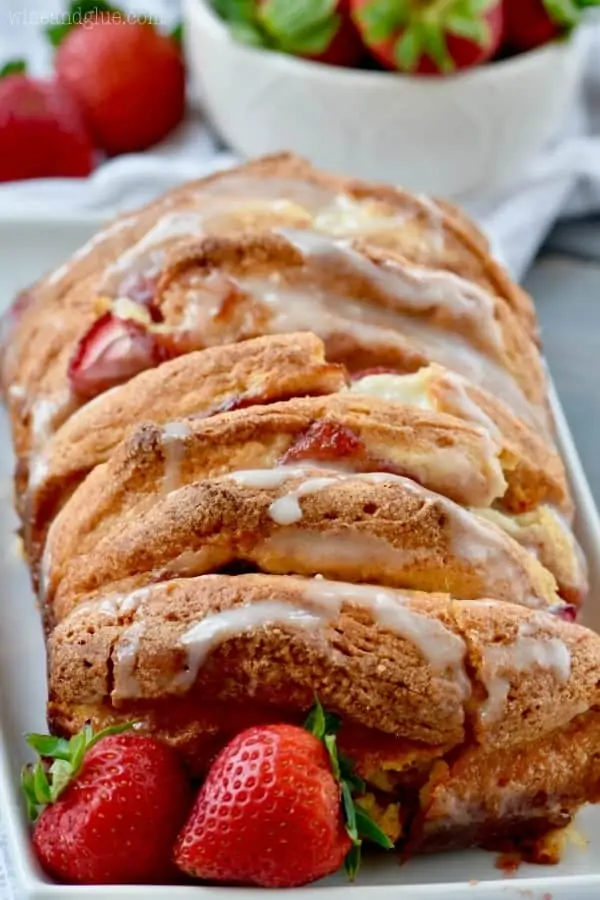 On a white rectangular plate with strawberries on the side, the Strawberries and Cream Pull Apart Bread has a beautiful golden brown crust with white icing on top. 