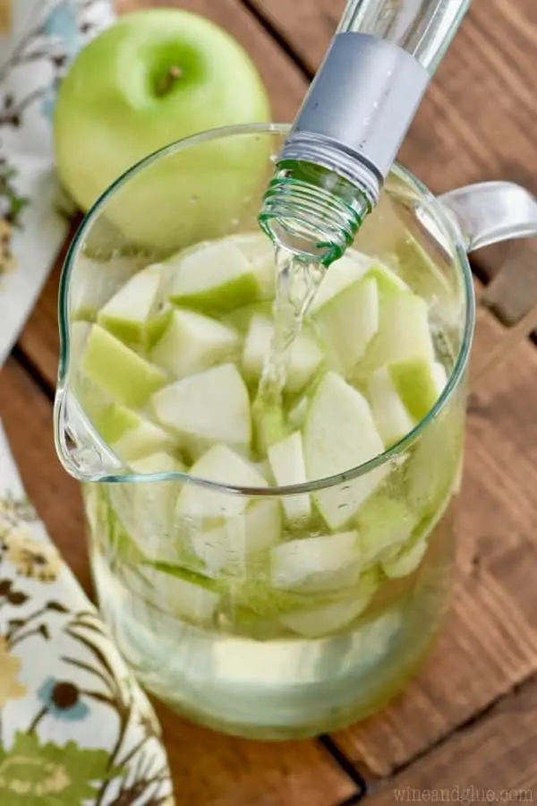 In a large glass pitcher, white wine is being poured on top of sliced green apples. 