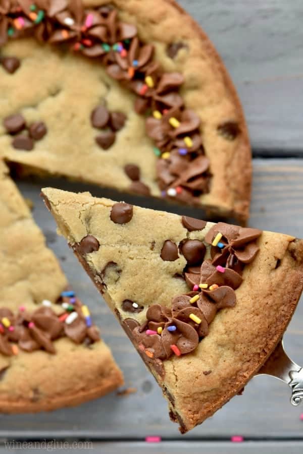 A slice of the Chocolate Chip Cookie Cake is being lifted up. There is a small border of chocolate frosting and rainbow sprinkles on the cookie cake. 