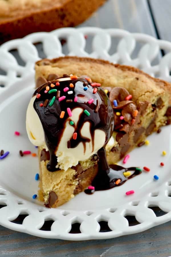 On a white plate, there is a slice of the Chocolate Chip Cookie Cake with a scoop of ice cream, chocolate syrup, chocolate frosting (as a border), and rainbow sprinkles 