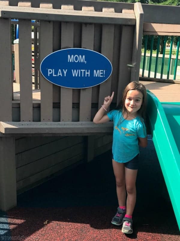 A little girl pointing at a sign in the park that says, "Mom, play with me!"