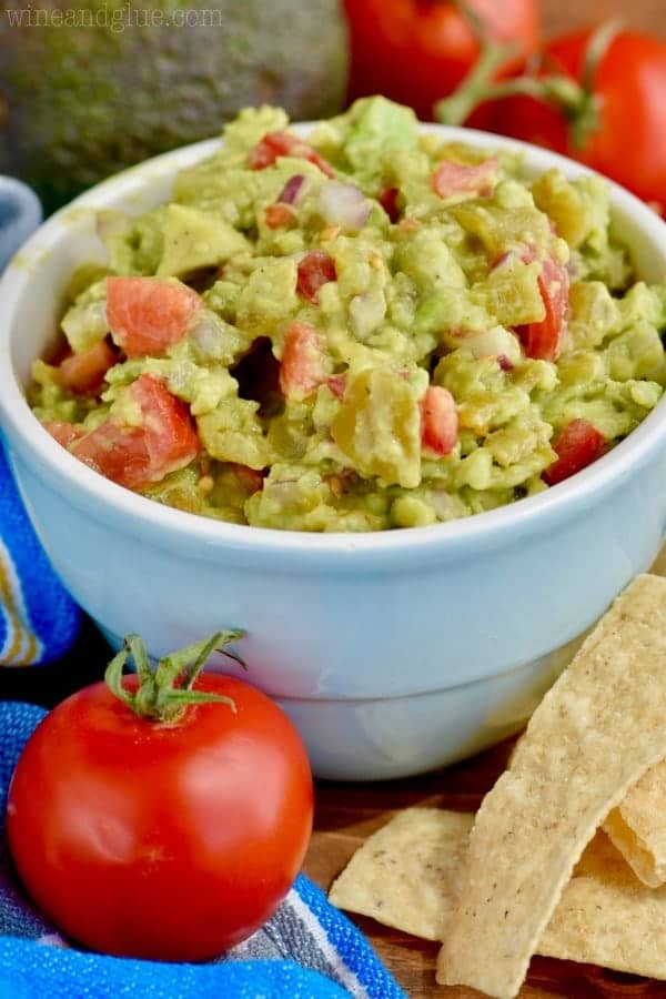 blue bowl filled with the best guacamole recipe with a tomato in front, avocados in the back, and chips on the side of it