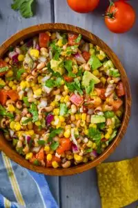 overhead view of a wood bowl on a blue wood surface surrounded by tomatoes and chips and with cowboy caviar inside