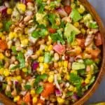 overhead view of a wood bowl on a blue wood surface surrounded by tomatoes and chips and with cowboy caviar inside