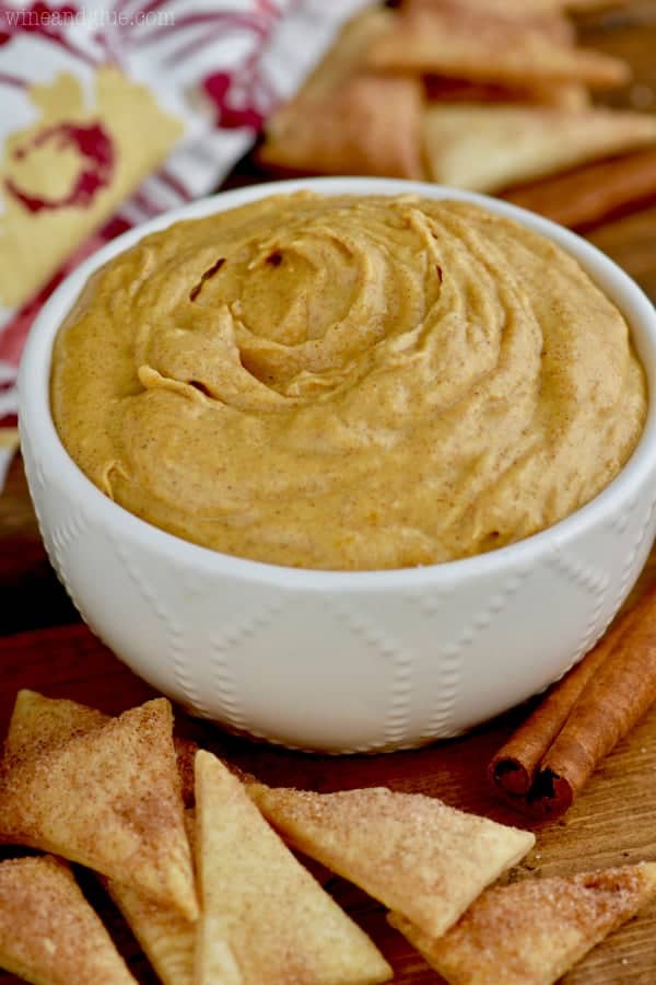 In a white bowl, the pumpkin pie dip has a golden brown hew with specks of cinnamon – surrounding the bowl are triangular cinnamon sugar pie crust. 