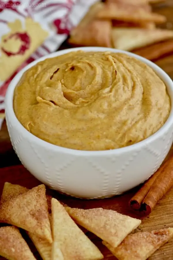 In a white bowl, the pumpkin pie dip has a golden brown hew with specks of cinnamon-- surrounding the bowl are triangular cinnamon sugar pie crust. 