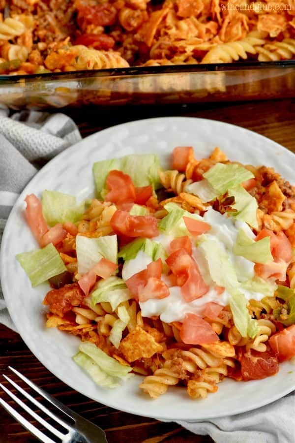 On a white plate is the Pasta Taco Casserole topped with sour cream, diced tomatoes, and lettuce. 