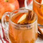 clear mug full of crockpot spiced apple cider with anise seed pods floating in it, apple slices and two cinnamon sticks