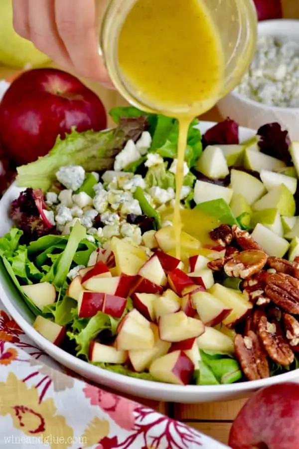 A salad with apples, blue cheese, pecans, and salad greens are being topped with a honey mustard dressing. 
