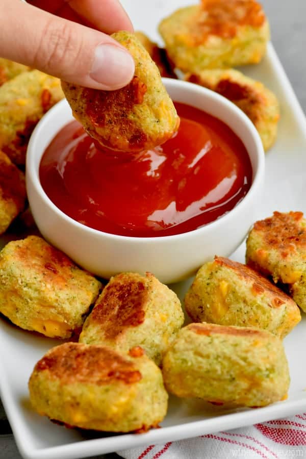 On a white plate, a small bowl of ketchup is surrounded by Broccoli Cheddar Cauliflower Tator Tots. One of the Broccoli Cheddar Cauliflower Tator Tot is being dipped into the ketchup. 