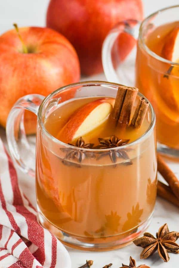 clear mug full of crockpot spiced apple cider with anise seed pods floating in it, apple slices and two cinnamon sticks