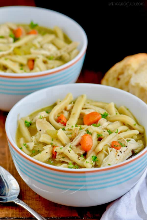 In a white bowl with blue stripes, the Homemade Chicken Noodle Soup has thick noodles and topped with minced parsley. 