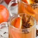 clear mug full of crockpot spiced apple cider with anise seed pods floating in it, apple slices and two cinnamon sticks