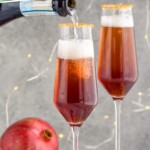 Pinterest graphic for Christmas Mimosa. Text says "the best christmas mimosa simplejoy.com" Image shows bottle of champagne pouring into a glass of Christmas mimosa ingredients with glass of Christmas mimosa and a pomegranate sitting in background