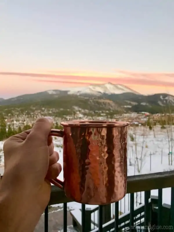 A woman holding a copper mug, and in the background are the beautiful Colorado Mountains. 