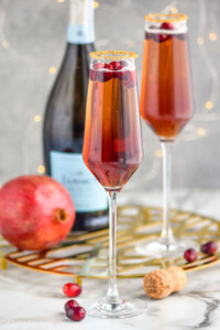 two champagne flutes of Christmas Mimosa with cranberries, pomegranate and bottle of champagne in background