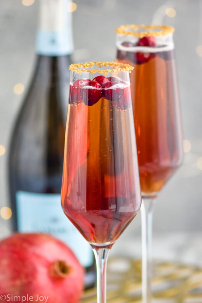 Two glasses of Cranberry mimosa cocktails with bottle of champagne and pomegranate sitting in background