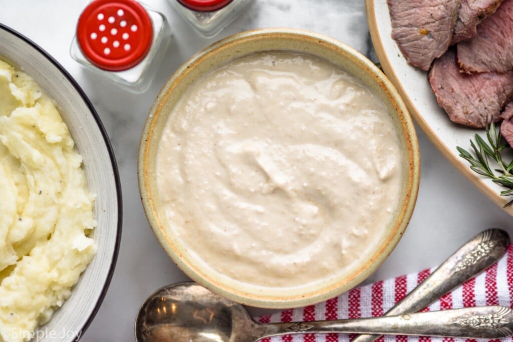 Overhead of bowl of homemade horseradish sauce with bowl of mashed potatoes and plate of prime rib sitting beside