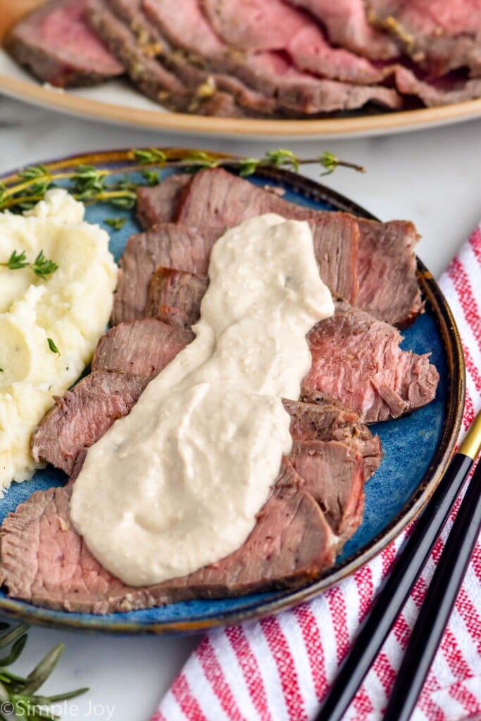plate of prime rib topped with horseradish sauce with mashed potatoes on on the side. Platter of prime rib for serving sitting in background.