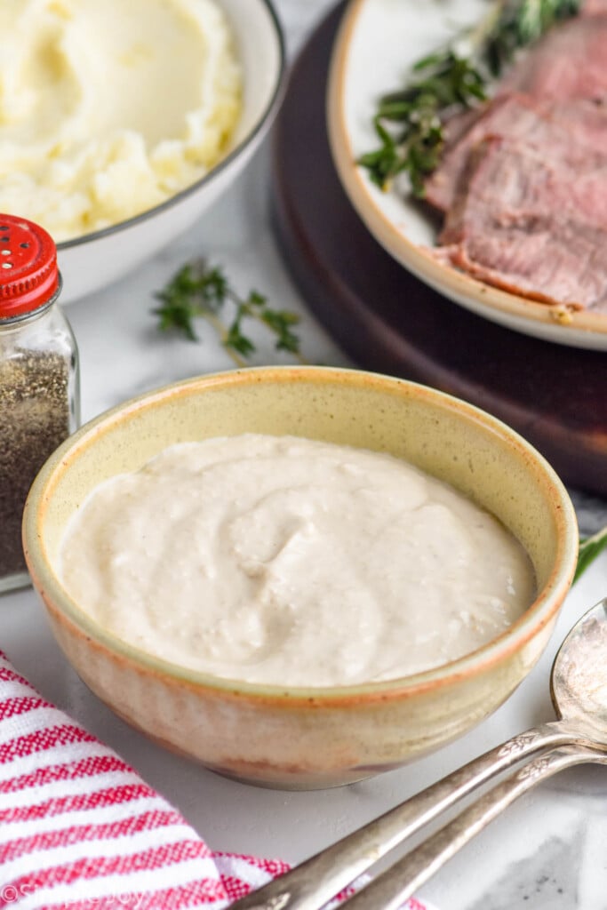 bowl of homemade horseradish sauce recipe with bowl of mashed potatoes and platter of prime rib in background for serving.