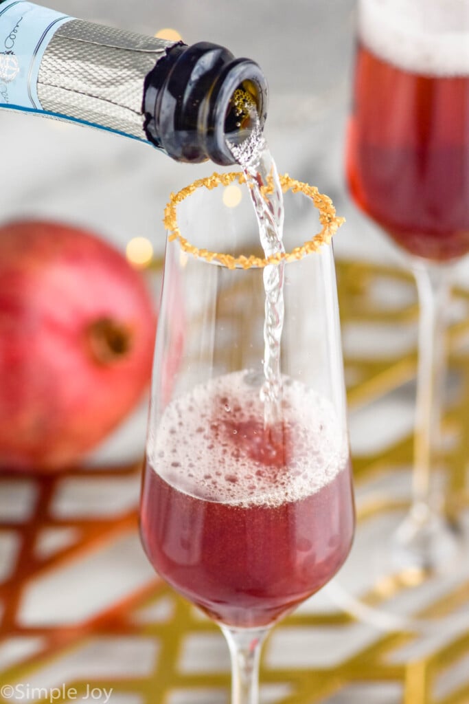 bottle of champagne pouring into a champagne flute to make a Christmas Mimosa. Glass of Christmas mimosa ingredients and pomegranate sitting in background