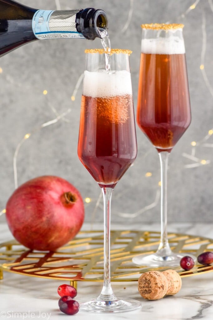 bottle of champagne pouring into a champagne flute to make a Christmas Mimosa. Glass of Christmas mimosa ingredients and pomegranate sitting in background