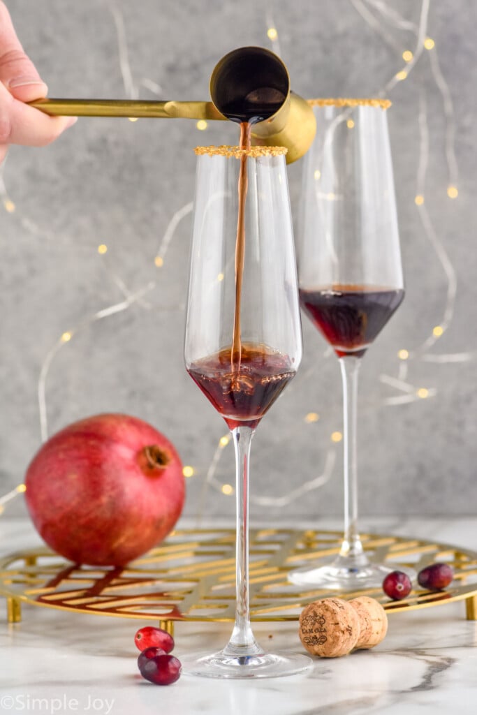 man's hang pouring cocktail jigger of pomegranate liqueur into a champagne flute to make a christmas mimosa. Glass of Christmas mimosa ingredients and pomegranate sitting in background