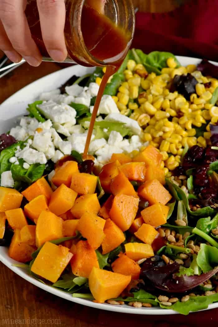 On a white plate, the Roasted Fall Salad with beautiful colors from cranberries, feta cheese, corn, greens, and butternut squash has the Maple Vinaigrette Dressing being poured on top. 