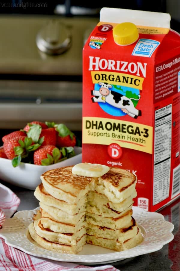 A stack of pancakes with a little slice cutout and a slice of butter on top. In the background, there are a bowl of Strawberries and carton of Milk. 