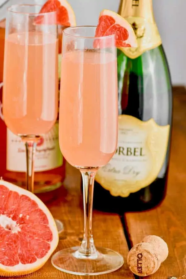 two champagne glasses filled with grapefruit champagne cocktail garnished with small wedges of fresh grapefruit, a half of a grapefruit in the background and a bottle of champagne