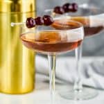 two long champagne coupe glasses filled with a Manhattan drink recipe, garnished with three cherries each, and a gold cocktail shaker in the background - 7
