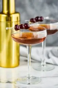 two long champagne coupe glasses filled with a Manhattan drink recipe, garnished with three cherries each, and a gold cocktail shaker in the background