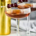 two long champagne coupe glasses filled with a Manhattan drink recipe, garnished with three cherries each, and a gold cocktail shaker in the background