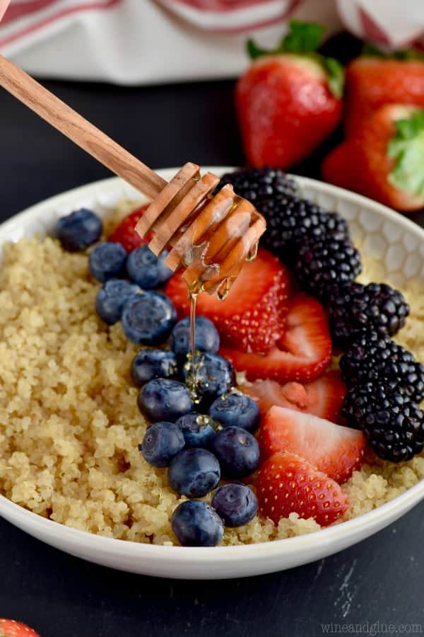 Honey is being drizzled on the bowl of Quinoa with blue berries, cut strawberries, and black berries. 