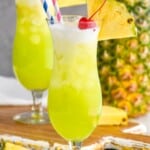 a hurricane glass filled with a midori splice garnished with a cherry and a pineapple wedge, some cream floating on top